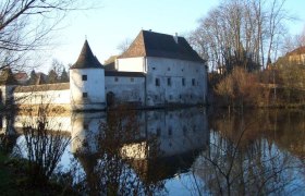 A historic building with towers is reflected in a calm body of water surrounded by bare trees.