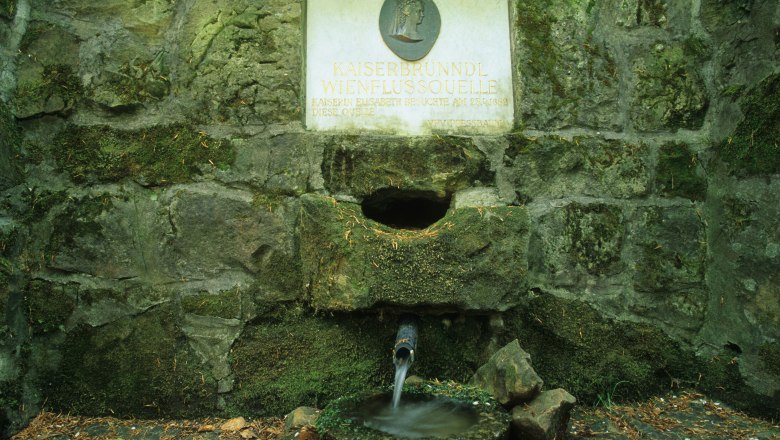 Stone fountain with inscription 'Kaiserbründl Wienflussquelle'. Water flows from a pipe into a bowl.