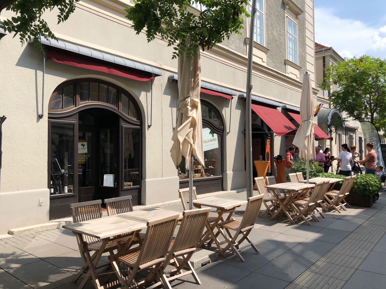 Außenbereich eines Cafés mit Holztischen und Stühlen, roter Markise und Menschen im Hintergrund.