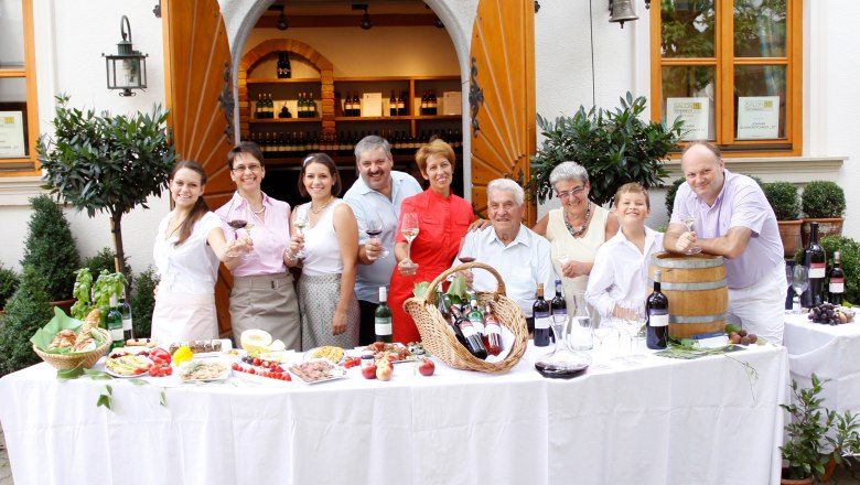 A group of people stand in front of a building with open wooden doors, smiling and holding glasses of wine. In front of them is a table with wine and food.