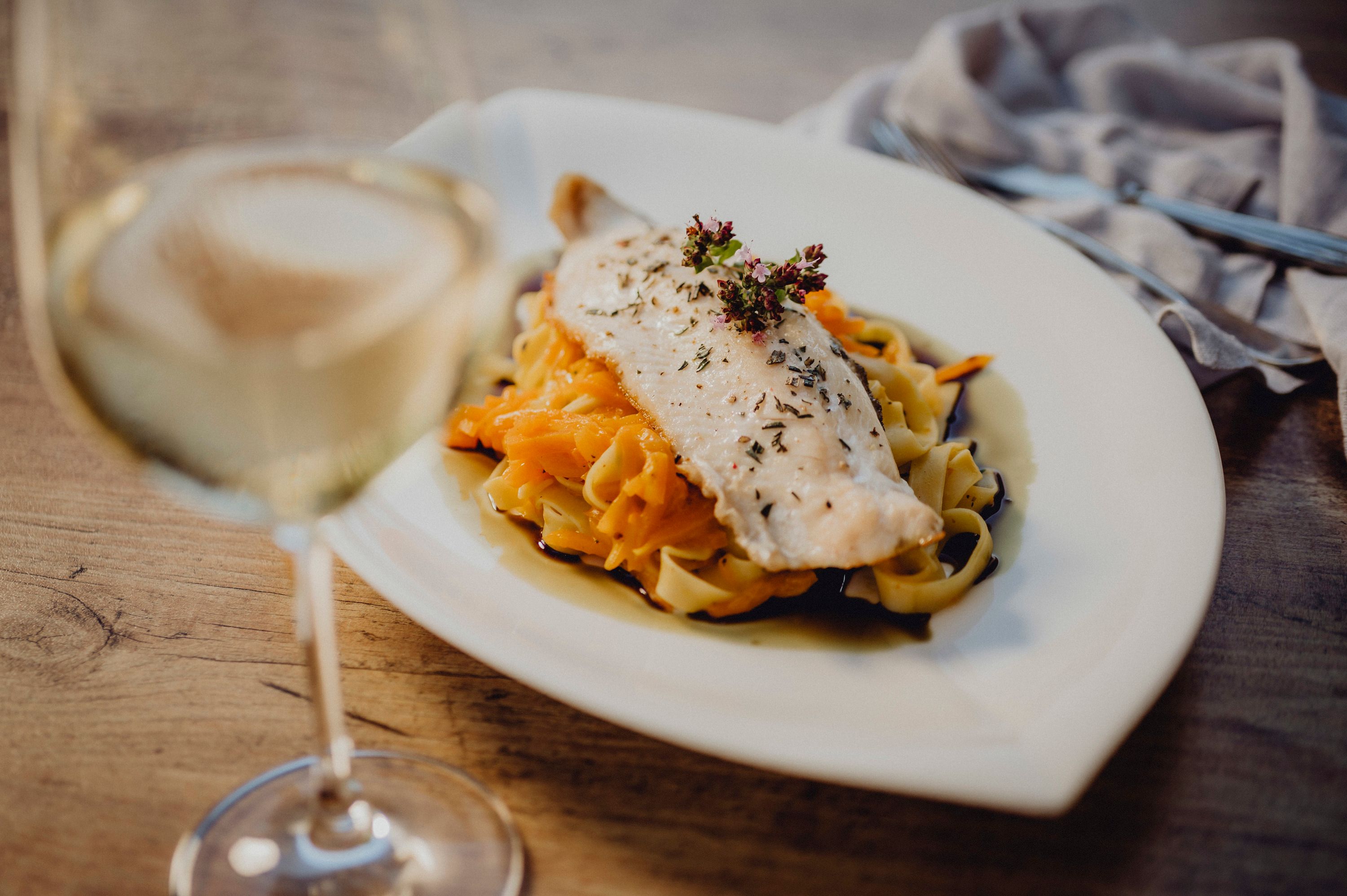 Ein Teller mit Fischfilet auf Nudeln und Gemüse, daneben ein Glas Weißwein.