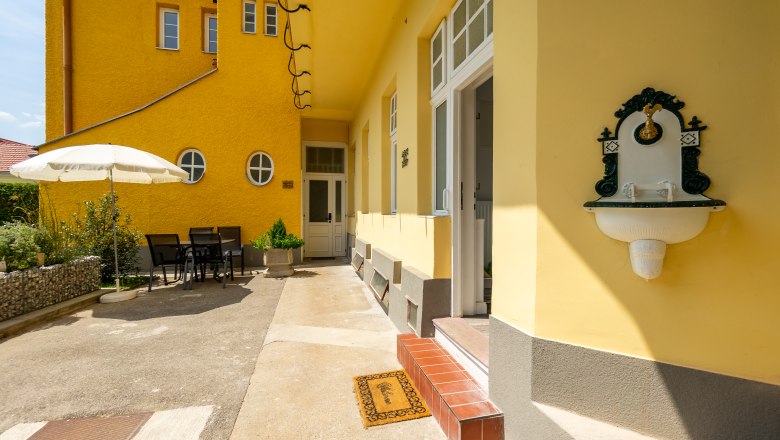 Gelbes Gebäude mit Terrasse, Tisch und Stühlen, Sonnenschirm und Wandbrunnen.