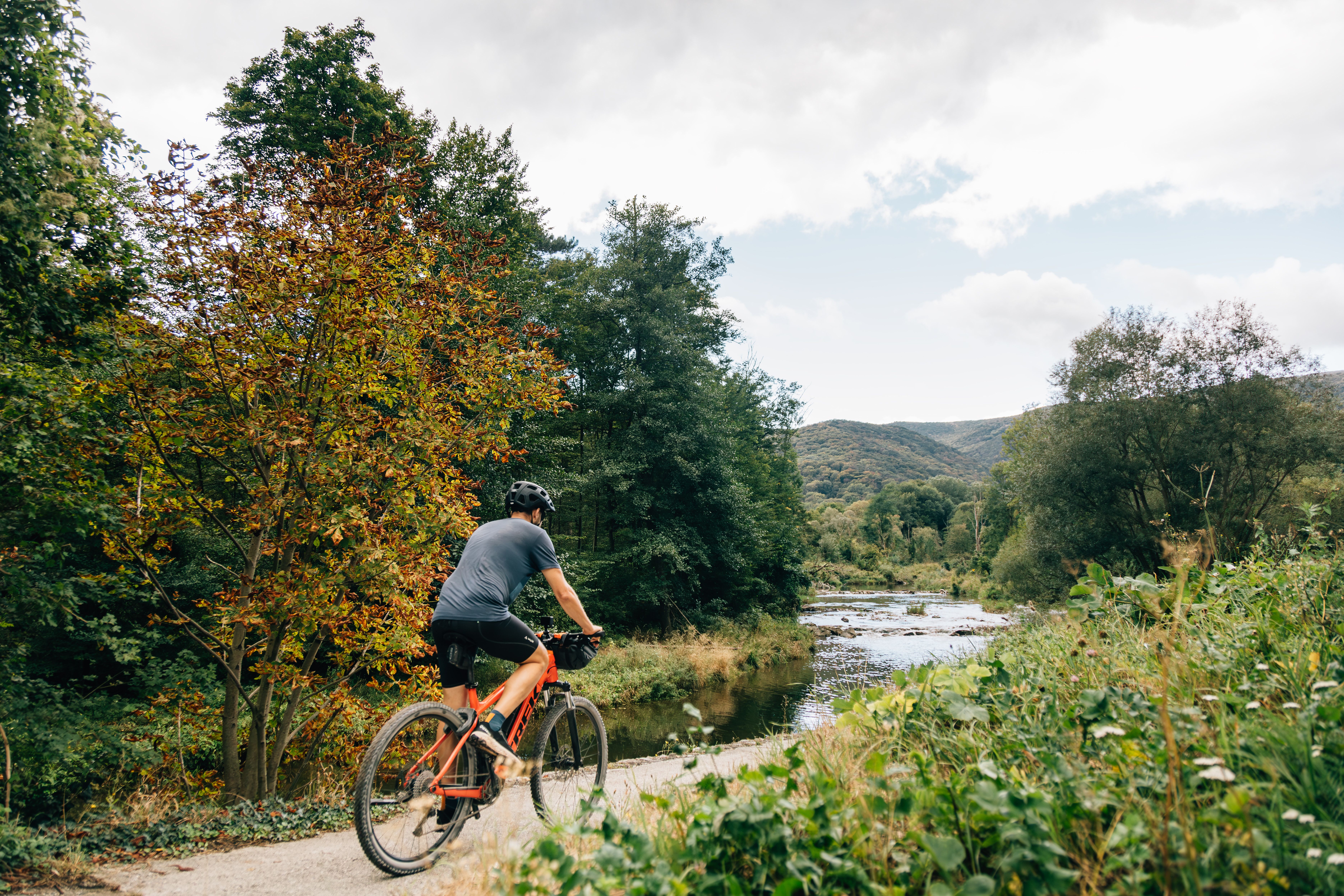 Ein Radfahrer genießt die frische Luft und die malerische Landschaft entlang eines ruhigen Flusses. Umgeben von üppigem Grün und bunten Herbstblättern, lädt die Umgebung zu einer entspannenden Auszeit in der Natur ein.