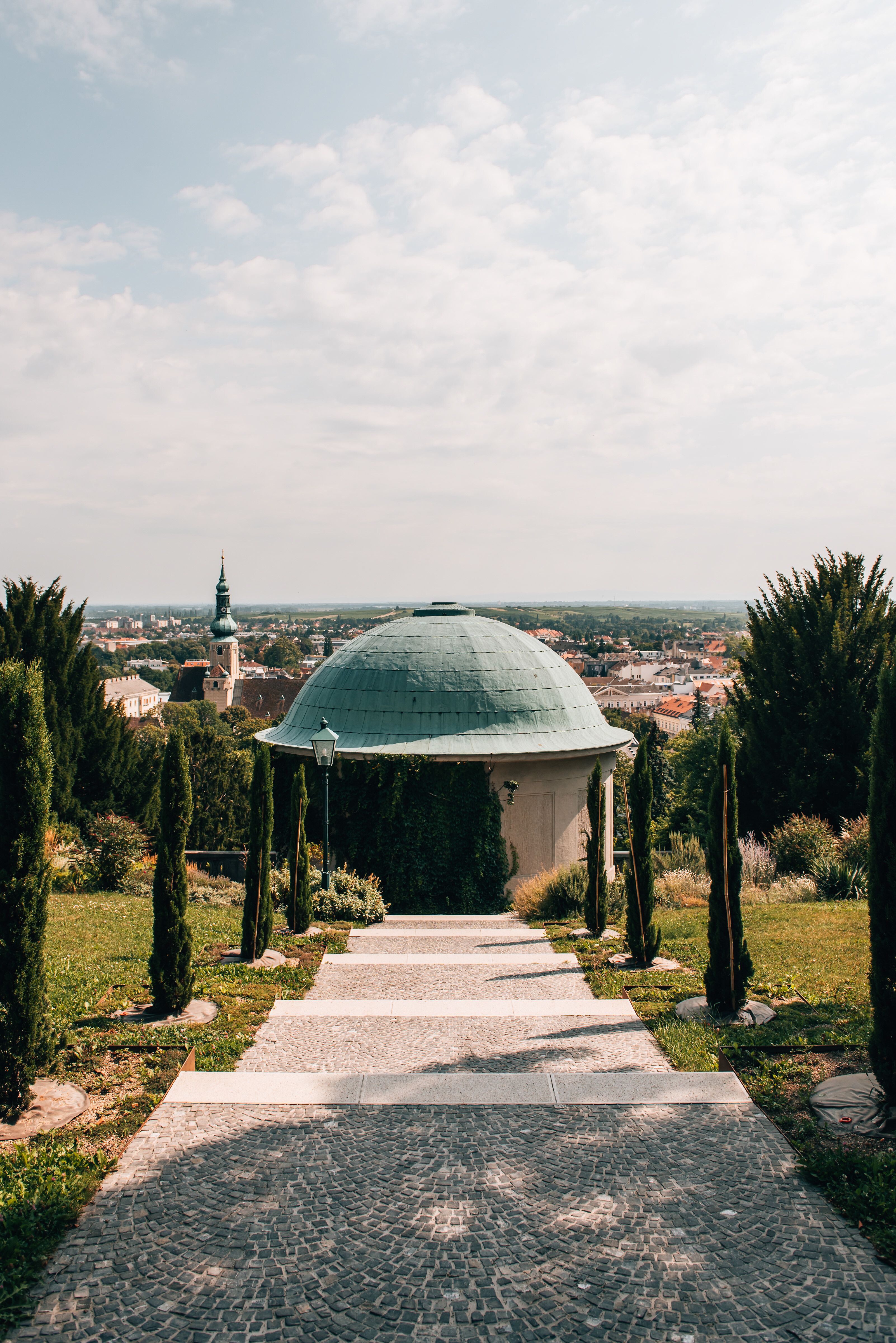 Ein sanfter Weg führt durch den üppigen Park, gesäumt von eleganten Zypressen, die Schatten spenden. In der Ferne erhebt sich eine charmante Kuppel, die den Blick auf die malerische Landschaft freigibt. Hier, wo die Natur und die Architektur harmonisch verschmelzen, lädt die Sommerfrische zum Verweilen und Entspannen ein.