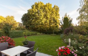 Ein gepflegter Garten mit Rasen, Blumenbeeten und Gartenmöbeln auf einer Terrasse.