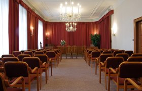 An elegant wedding hall with red curtains, chandeliers and rows of chairs.