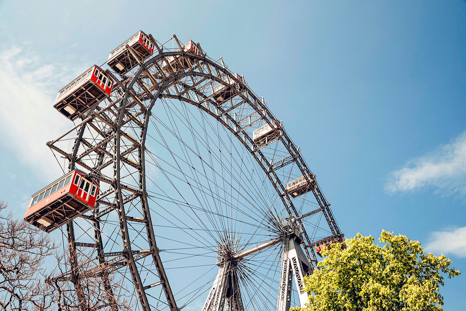 Das Wiener Riesenrad vor blauem Himmel mit grünen Bäumen im Vordergrund.