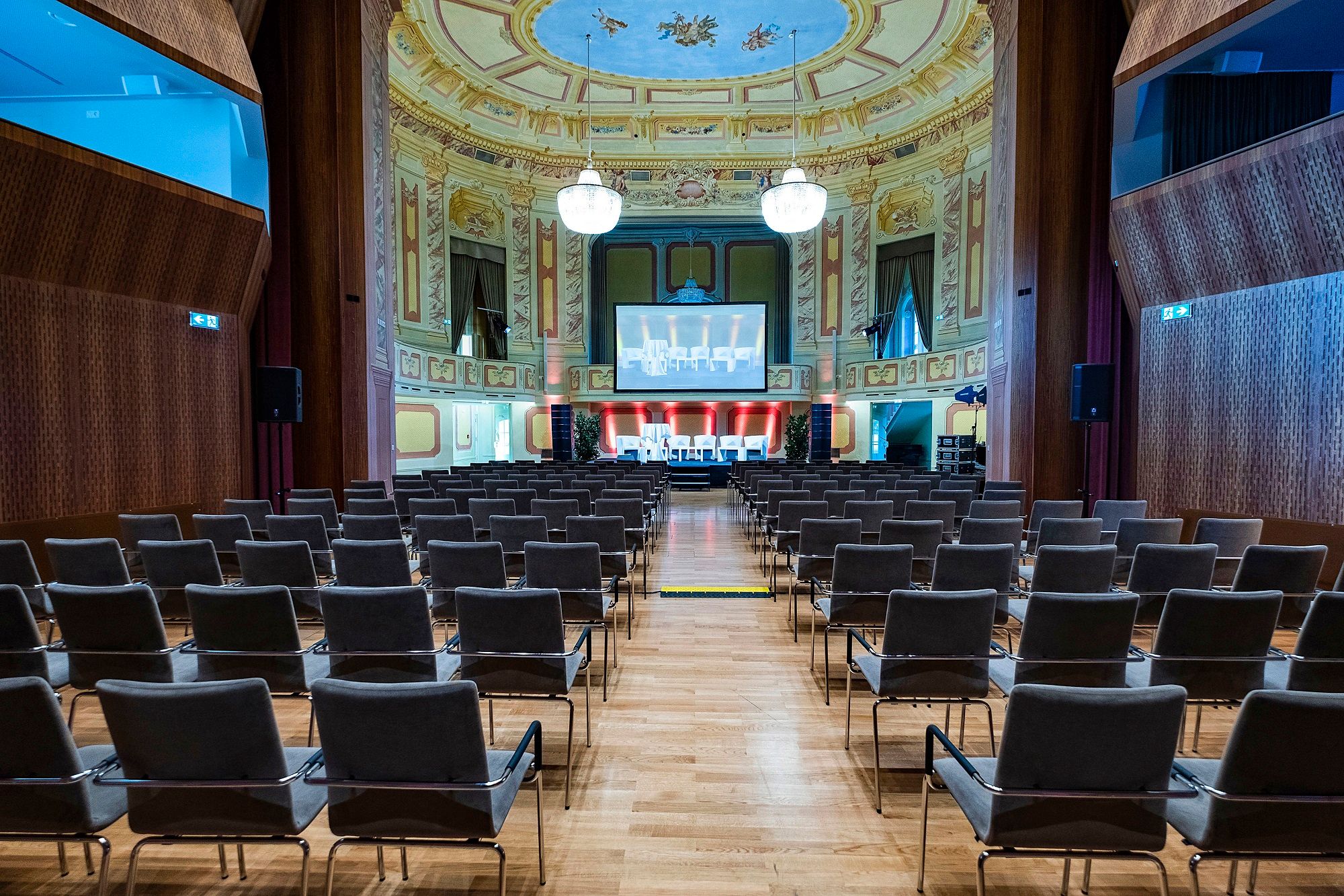 Innenansicht eines Theatersaals mit Stuhlreihen, einer Bühne und einem großen Bildschirm.