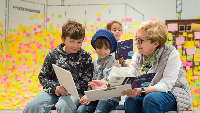 An elderly woman and two children sit together and read in a room with colorful sticky notes on the wall.