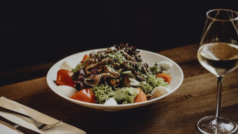 Ein Teller mit gemischtem Salat, Tomaten, Eiern und Fleischstreifen auf einem Holztisch, daneben ein Glas Weißwein.