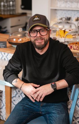 Michael Kulhanek sitzt entspannt in seinem Lokal "Kulhanek's Kaffee & Laden". Eine Reihe köstlicher Torten schmückt dahinter die Theke . Ein Ort, wo Genuss im Licht badet und die süßen Versuchungen voller Erwartung strahlen.