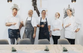 Five people dressed as chefs are laughing and holding glasses of wine.