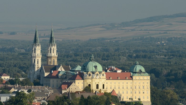 Luftaufnahme des Stifts Klosterneuburg mit zwei Türmen und einer Kuppel, umgeben von Wald und Feldern.