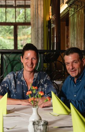 Brigitte und Connie Blanka, sitzen in ihrem gemütlichen Restaurant an einem Tisch und lächeln in die Kamera. Hinter ihnen reihen sich weitere Tische, während ein ausgestopfter Hirsch an der Wand thront und eine rustikale Kulisse schafft.