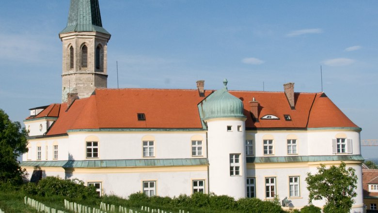 Gumpoldskirchen Castle, © Schloss Gumpoldskirchen Betriebs-GmbH Gumpoldskirchen Castle with its red roof and tower, surrounded by vineyards under a blue sky.