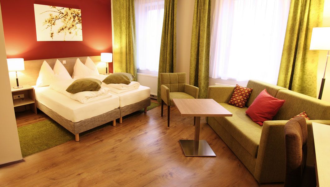 Gemütliches Hotelzimmer mit Doppelbett, grüner Couch, Tisch und Stuhl. Warme Farben und moderne Einrichtung.