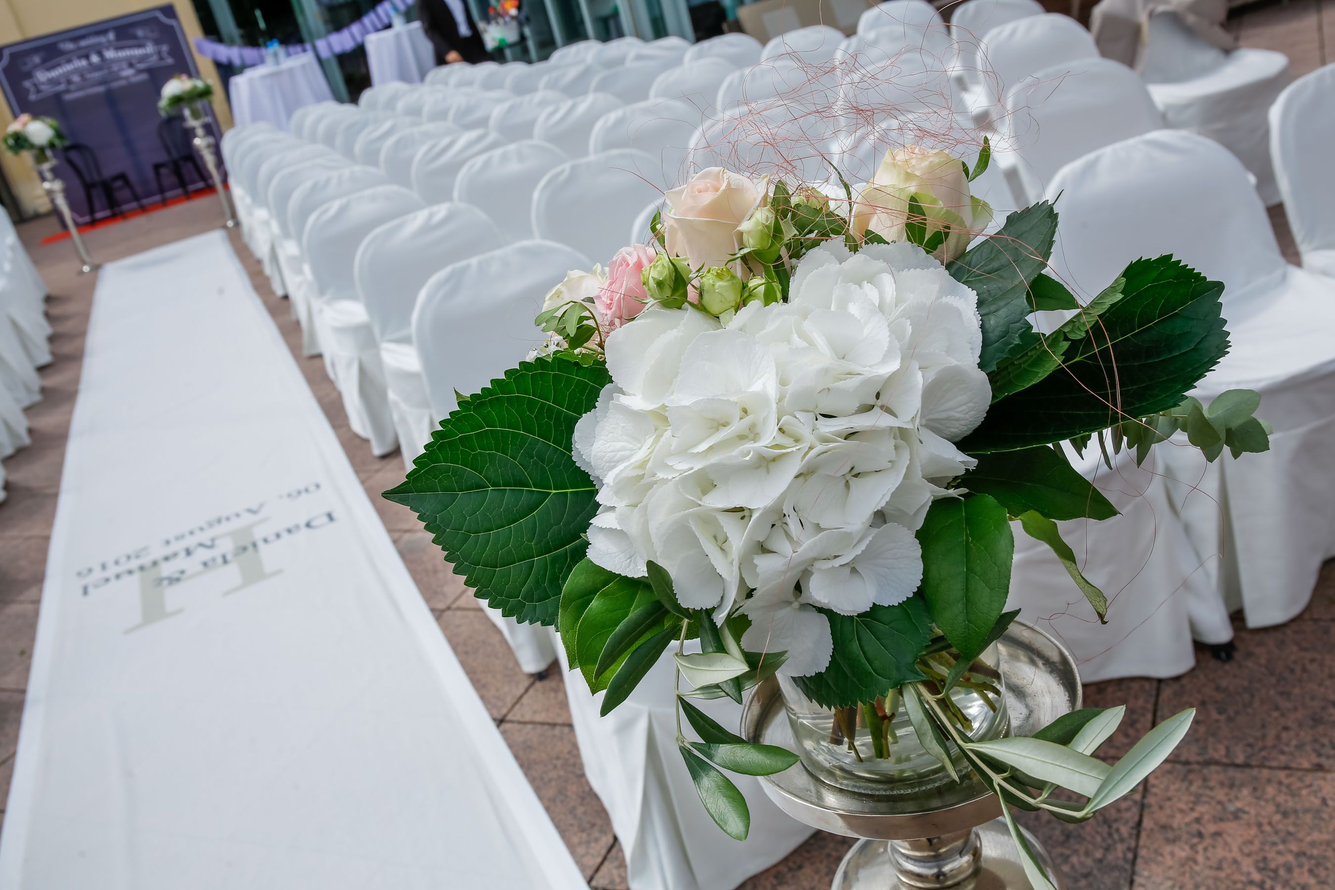 Mehrere mit festlichen weißen Hussen dekorierte Stühle sind vorbereitet für eine Zereomie, im Vordergrund steht ein weißer Blumenstrauß.