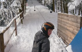 A child wearing a helmet stands on skis at a drag lift in a snowy forest.