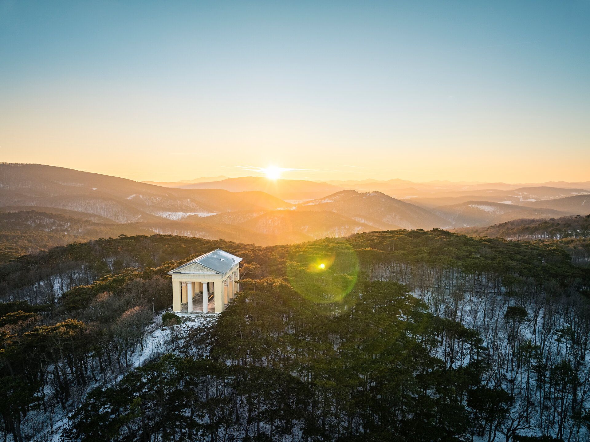 Drohnenaufnahme des Husarentempels im winterlichen Wienerwald, mit verschneiten Baumkronen und Blick über die hügelige Landschaft im warmen Licht des Sonnenaufgangs.