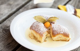 Drei Buchteln mit Puderzucker und Physalis auf Vanillesauce auf einem weißen Teller.