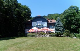 Ein großes Haus namens Rudolfshof mit Terrasse und Sonnenschirmen, umgeben von Bäumen und einer Wiese.