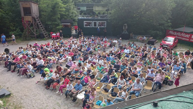 An audience sits outside on chairs in a quarry, surrounded by trees and a red car with Media Markt advertising.