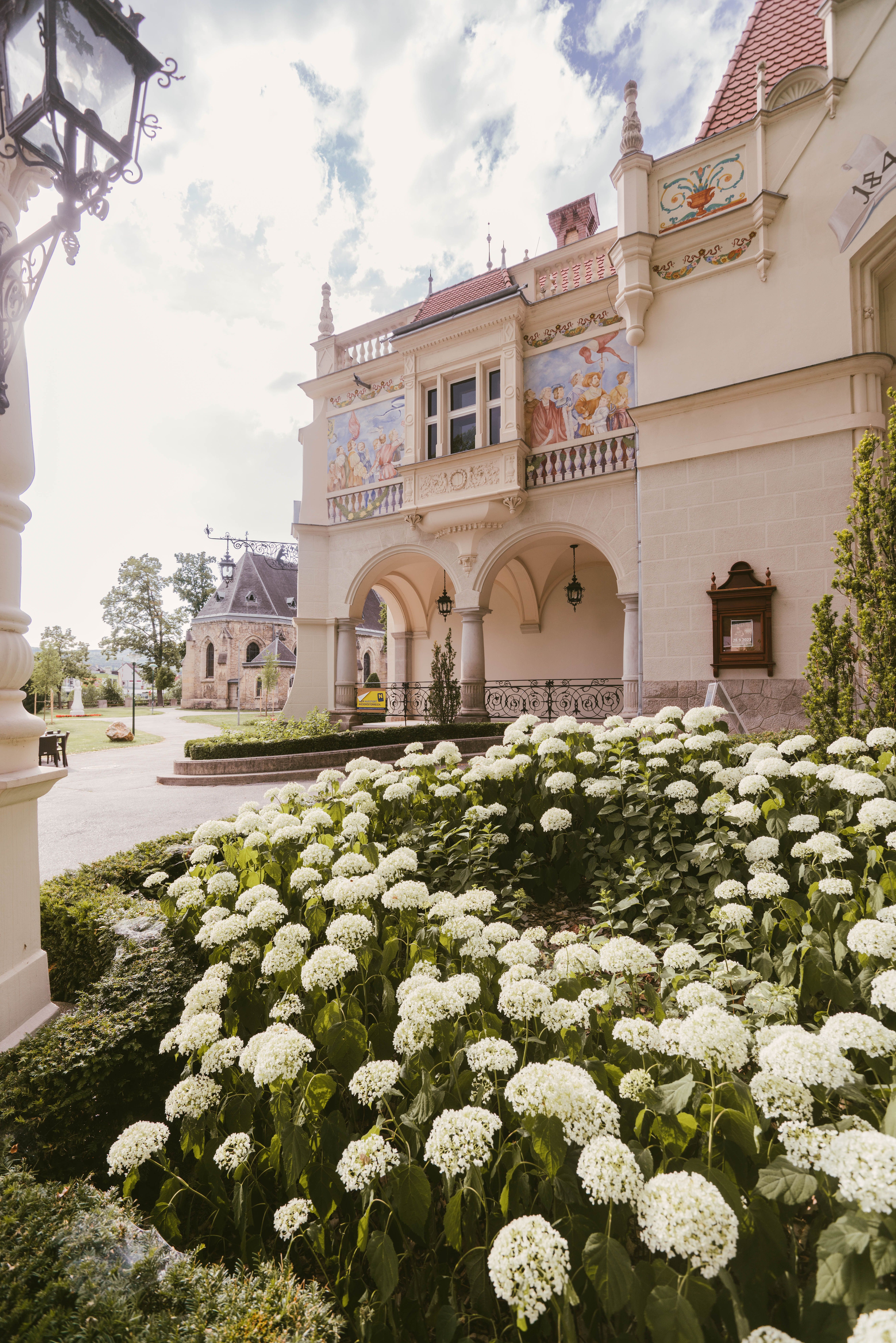 Umgeben von üppigen, blühenden Hortensien entfaltet sich die majestätische Fassade des Stadttheaters. Die kunstvollen Verzierungen und die harmonische Architektur laden dazu ein, in die kulturelle Vielfalt der Region einzutauchen. Ein Ort, an dem Kunst und Natur in perfekter Symbiose verschmelzen.