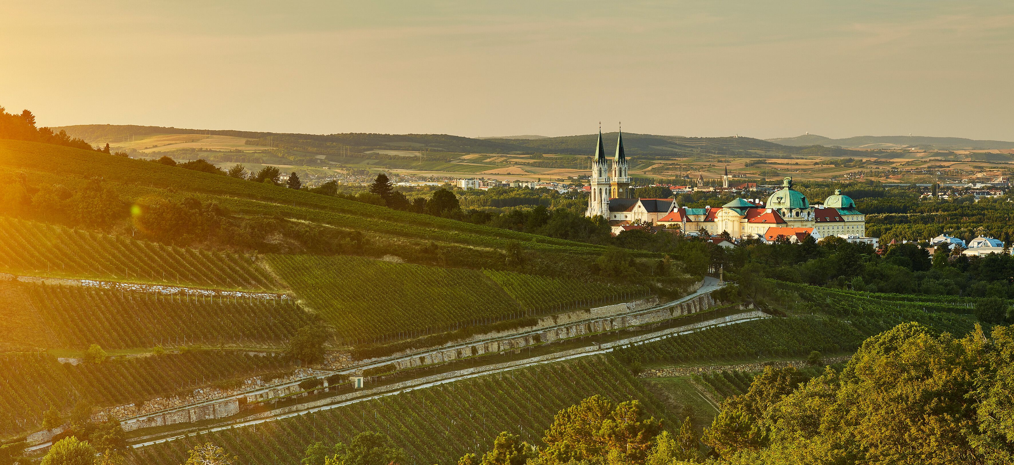 Die sanften Hügel des Wienerwaldes umarmen das majestätische Stift Klosterneuburg, während die goldenen Strahlen der Abendsonne die Weinreben in ein warmes Licht tauchen. Hier, wo Natur und Kultur harmonisch verschmelzen, lädt die malerische Landschaft zu unvergesslichen Erlebnissen ein.