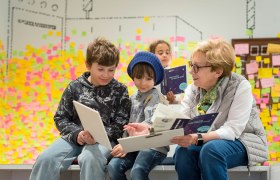 Eine &auml;ltere Frau und zwei Kinder sitzen zusammen und lesen in einem Raum mit bunten Haftnotizen an der Wand.