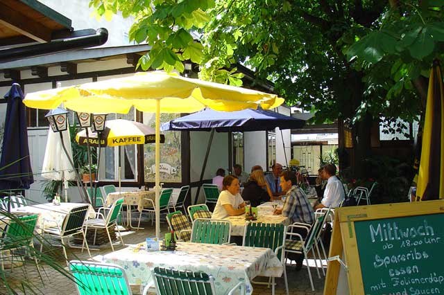 Ein gemütlicher Gastgarten mit Tischen, Stühlen und Sonnenschirmen, umgeben von Bäumen. Menschen sitzen und unterhalten sich.